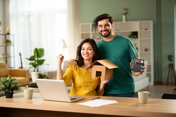 Happy Indian asian young couple working on finance using laptop, holding money fan and house model
