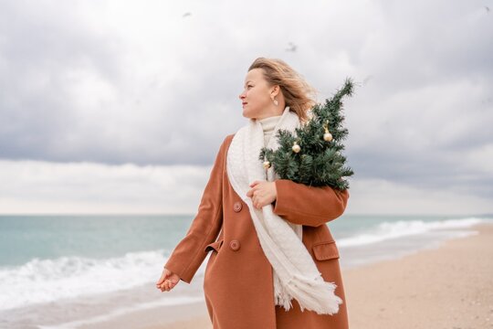 Blond woman Christmas tree sea. Christmas portrait of a happy woman walking along the beach and holding a Christmas tree on her shoulder. She is wearing a brown coat and a white suit.