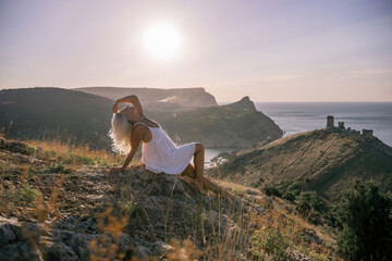 A woman is sitting on a hillside overlooking the ocean. She is wearing a white dress and has blonde hair. The scene is serene and peaceful, with the ocean in the background.