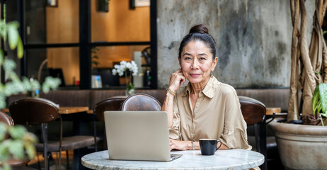 An elderly woman of 60  70 years old is sitting in a modern cafe with a laptop. Senior happy business woman using pc technologies, working, chatting, spending time in social media internet, ecommerce.