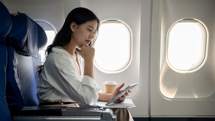 An attractive, hard-working Asian businesswoman is working on her tablet while traveling by plane. - obrazy, fototapety, plakaty