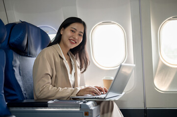 An attractive Asian businesswoman is on a flight, sitting at a window seat with her laptop.