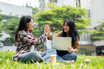 Two cheerful college students are giving each other a high five while sitting on the grass in a park