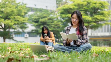 A focused Asian female student is reading a book while sitting on the grass in a campus park.
