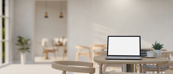 A white-screen laptop computer mockup on a table in a spacious minimalist restaurant or coffee shop.