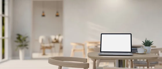 Foto op Plexiglas A white-screen laptop computer mockup on a table in a spacious minimalist restaurant or coffee shop. © bongkarn