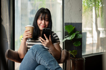 A happy Asian female is relaxing in a coffee shop, using her smartphone and enjoying her coffee. - 776920353