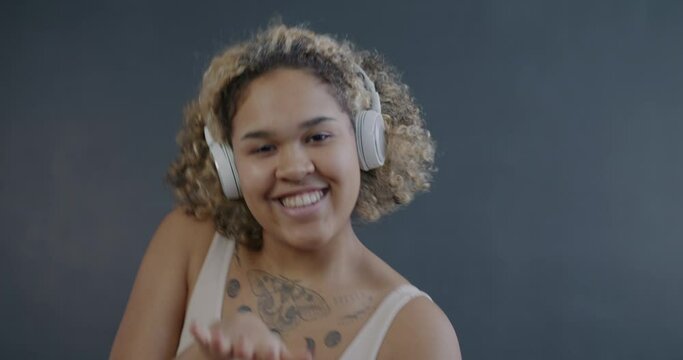 Overjoyed African American woman wearing headphones dancing enjoying music looking at camera on grey background. Emotion and modern lifestyle concept.