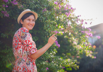 young asian woman stands in sunny green park, admiring blooming bougainvillea bush. The flower's bright colors are beautiful contrast to park's lush greenery.