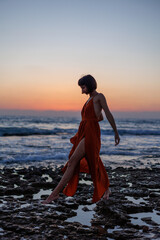young woman in a summer dress walks along the seashore during sunset and looks at the sea. - 776949555
