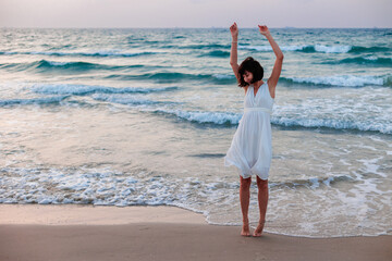 Happy woman in a white dress enjoys relaxing on the beach, walking along the sandy shore at sunset. - 776952109