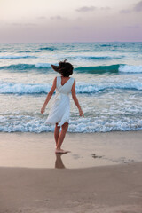 Happy woman in a white dress enjoys relaxing on the beach, walking along the sandy shore at sunset. - 776952527
