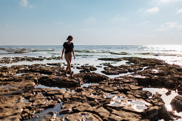 A teenage girl walks alone along the water line on a sand and stone beach during summer vacation. \ - 776956954