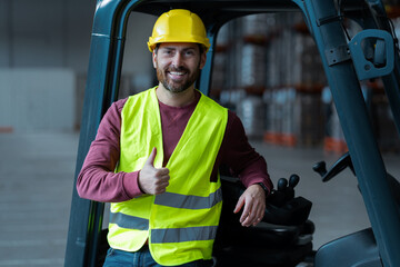 Bearded man, driver showing thumbs up, wearing helmet, standing near forklift, looking at camera