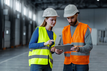 Attractive man and woman, engineers wearing protective workwear and hard hat using digital tablet