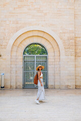 Back view of a hipster girl traveler in a hat and backpack traveling alone through the sights of Israel. - 776958561