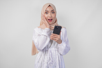 Naklejka premium Excited Asian woman wearing hijab and white dress smiling happily while holding smartphone, standing over isolated white background. Ramadan and Eid Mubarak concept