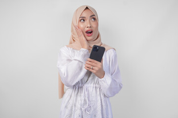 Fototapeta premium Excited Asian woman wearing hijab and white dress smiling happily while holding smartphone, standing over isolated white background. Ramadan and Eid Mubarak concept