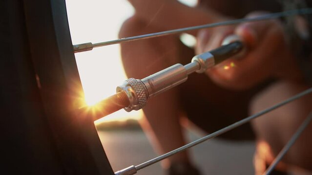 Close up cyclist inflates flat tire by compact hand pump with retractable hose with Schrader inflator chuck connector for valve stem at sunset, punctured wheel, broken bicycle out of town outside city