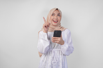 Thoughtful young Asian Muslim woman wearing white dress and hijab holding phone while thinking and finding idea with happy expression, isolated on white background. Ramadan and Eid Mubarak concept