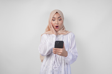 Naklejka premium Young Asian Muslim woman wearing white dress and hijab showing shocked expression on her face while holding smartphone, surprised after reading news or gossip, isolated by white background