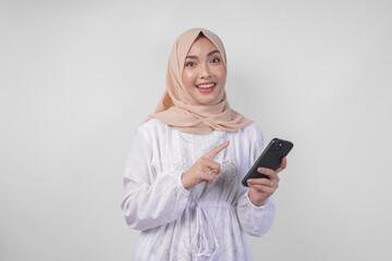 Excited Asian Muslim woman wearing white dress and hijab holding her smartphone while smiling cheerfully, isolated by white background. Ramadhan and Eid Mubarak concept