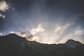 The rays of the setting sun shine from behind a mountain and a rocky ridge in the Fan Mountains in...