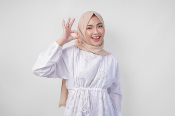 Fototapeta premium Portrait of Asian Muslim woman in white dress and hijab making an okay, good, nice sign gesture using fingers. Ramadan and Eid Mubarak concept