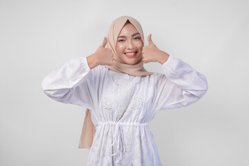 Fototapeta premium Attractive young Asian woman in white dress and hijab gesturing thumbs up for approval, good, excellent signs, isolated by white background