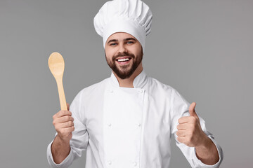 Happy young chef in uniform holding wooden spoon and showing thumb up on grey background