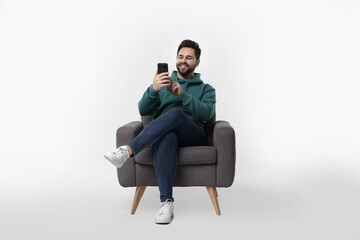Happy young man using smartphone in armchair on white background