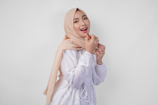 Beautiful Young Asian Muslim Woman Wearing White Dress And Hijab Putting On Makeup Applying Lipstick. Fashion And Cosmetics Beauty Concept