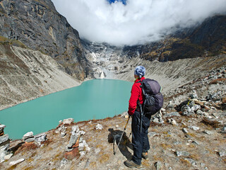 A hiker stands before a turquoise glacial lake surrounded by rugged mountains shrouded in clouds