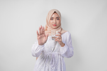 Serious young Asian Muslim woman wearing white dress and hijab showing her palm to make a stop...