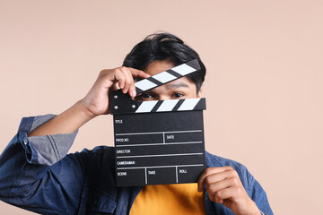 Young man holding clapperboard in front of his face, eyes peeking against beige background
