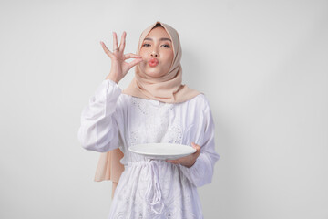 Happy young Asian Muslim woman in hijab holding an empty plate with copy space and making a...