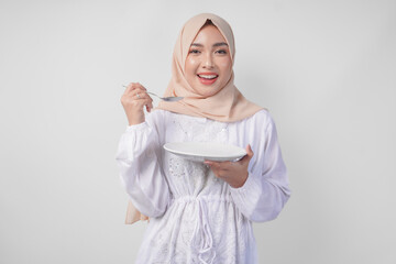 Hungry young Asian Muslim woman in hijab holding spoon and eating from an empty plate with copy space over. Ramadan concept
