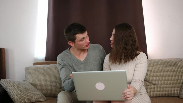 Young couple, a family, sits on the sofa, looks at the laptop screen, applies for an online business loan, opens their first business, and makes a joint decision to purchase commercial real estate