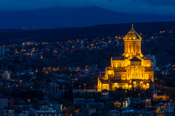 Night view of the historical district of Tbilisi from the mountain. Holy Trinity Cathedral of Tbilisi, Avlabari. Dark blue sky, orange lighting of the church. - obrazy, fototapety, plakaty