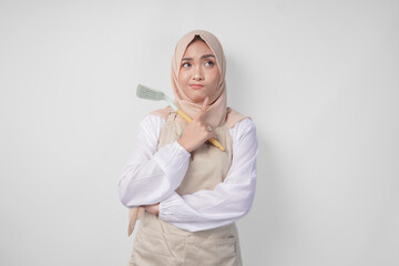 Thoughtful young Asian Muslim woman in hijab and cream apron holding spatula and kitchen cooking...