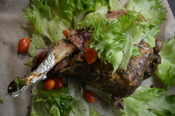 Eating, cooking delicious meat, sheep leg on the bone marinated and baked in the oven. A large piece of lamb arranged on brown parchment paper with green lettuce leaves and small cherry tomatoes.