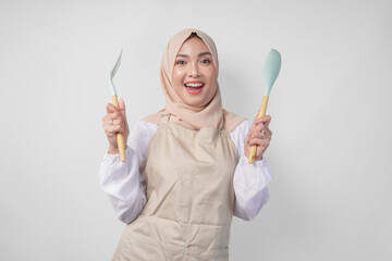 Excited young Asian Muslim woman in a veil hijab and cream apron smiling to the camera while...