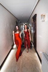 Two sexy women in red dresses ride on a golden luggage trolley in a hotel. Fashion shooting concept