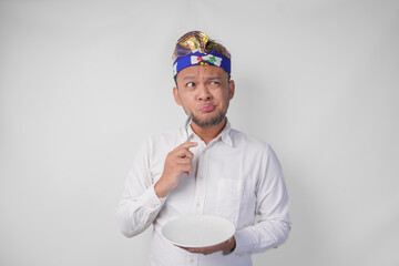 Thoughtful young Balinese man in white shirt and traditional headdress holding an empty plate with copy space and spoon while thinking what food to eat