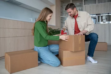 Fotobehang Happy couple packing cardboard box using sticky tape to move in new apartment together. Family wife husband prepare to relocate in flat house. Relocation mortgage, life changes, buying real estate. © DimaBerlin