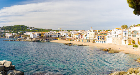 Fototapeta premium Amplia vista panorámica de la Playa del Canadell en Calella de Palafrugell en invierno, con edificios blancos a lo largo de la costa y cielo parcialmente nublado.