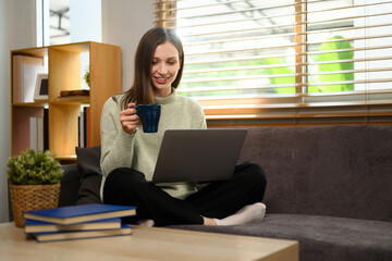 Beautiful caucasian lady drinking hot tea and using laptop in living room at home