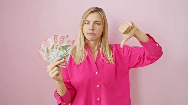 Angry blonde lass thumbs down at new zealand dollars, showing raw dislike over pink isolated background
