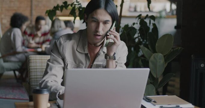 Asian freelancer speaking on mobile phone and working with laptop computer sitting at table in coworking office. Freelance job and communication concept.