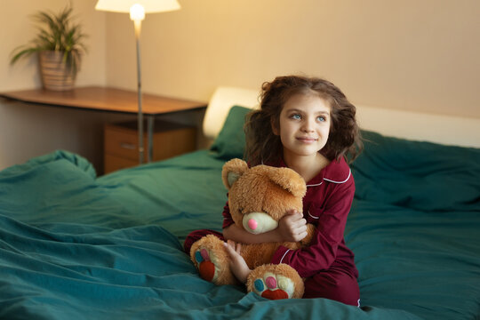 7 year old girl in red pyjamas sits in green bed with her teddy bear toy at home